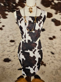 A black and white cow print dress displayed on a mannequin torso, styled with a gold chain necklace, set against a brown and white cowhide rug.