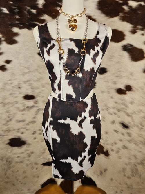 A black and white cow print dress displayed on a mannequin torso, styled with a gold chain necklace, set against a brown and white cowhide rug.