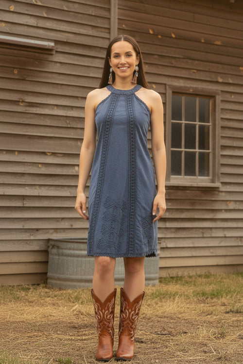 Woman wearing a blue dress and brown cowboy boots standing in front of a wooden building.
