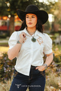 Woman wearing a white shirt with embroidery, black hat, and dark jeans in an outdoor setting.
