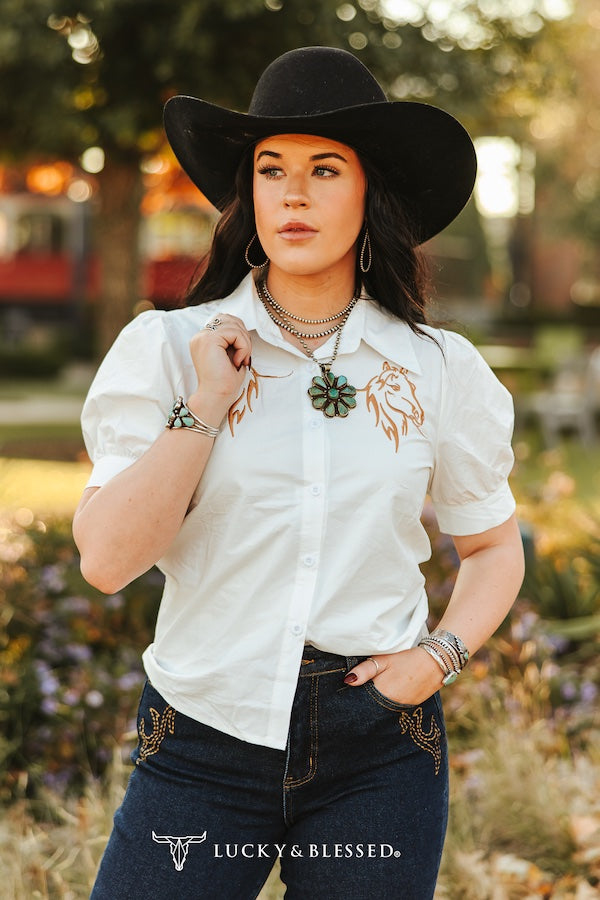 Woman wearing a white shirt with embroidery, black hat, and dark jeans in an outdoor setting.